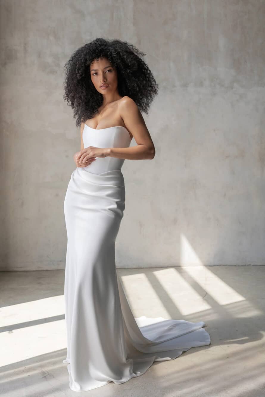 Alyssa Kristin Bridal, Kaia, a sculpted Mikado bodice that offers clean architectural lines, while a bias-cut satin skirt features pleating that accentuates the basque waist and continues effortlessly into a fluid silhouette at Love and Lace Bridal Salon in Orange, CA.