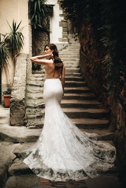 Zita by Anais Anette Bridal is a wedding dress that features a silhouette-enhancing silhouette crafted from light and stretchy crepe material with a soft V-neckline, thin straps, and an open low back with lace triangle side cutouts and a signature illusion train with an overblown floral detail available at Love and Lace Bridal Salon in Orange County, CA.