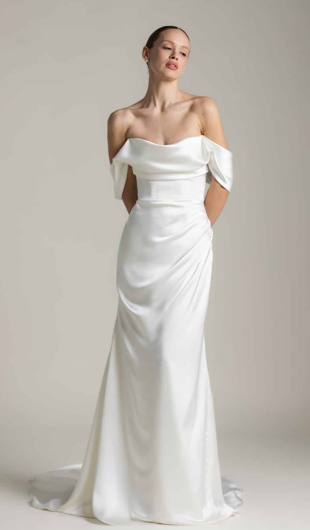 The Athena wedding dress by Aesling Bridal, available at Love and Lace Bridal Salon in Orange, CA, features a minimalist liquid-satin sculptural silhouette, a strapless plunging V-neckline, draped off-the-shoulder swag sleeves, and an 80-inch flared train.