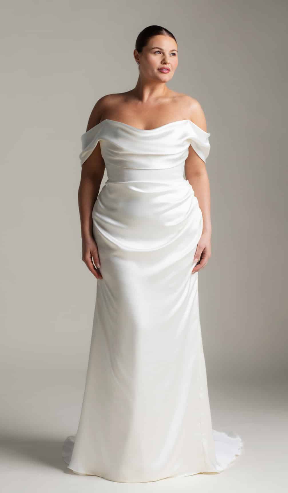 The Athena plus size wedding dress by Aesling Bridal, available at Love and Lace Bridal Salon in Orange, CA, features a minimalist liquid-satin sculptural silhouette, a strapless plunging V-neckline, draped off-the-shoulder swag sleeves, and an 80-inch flared train.
